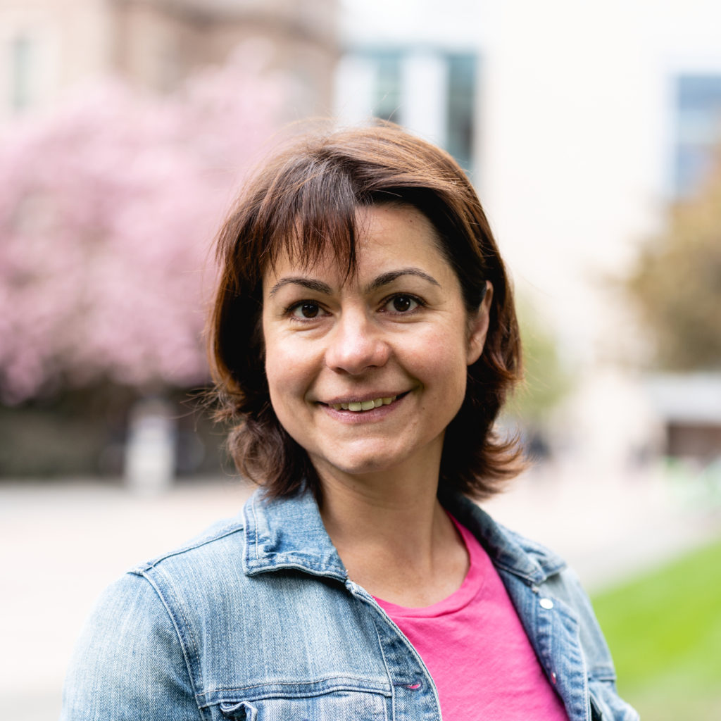 A smiling photo of Elena Mohova, Hall Health. Elena has medium brown hair and is wearing a pink t shirt with a denim jacket.