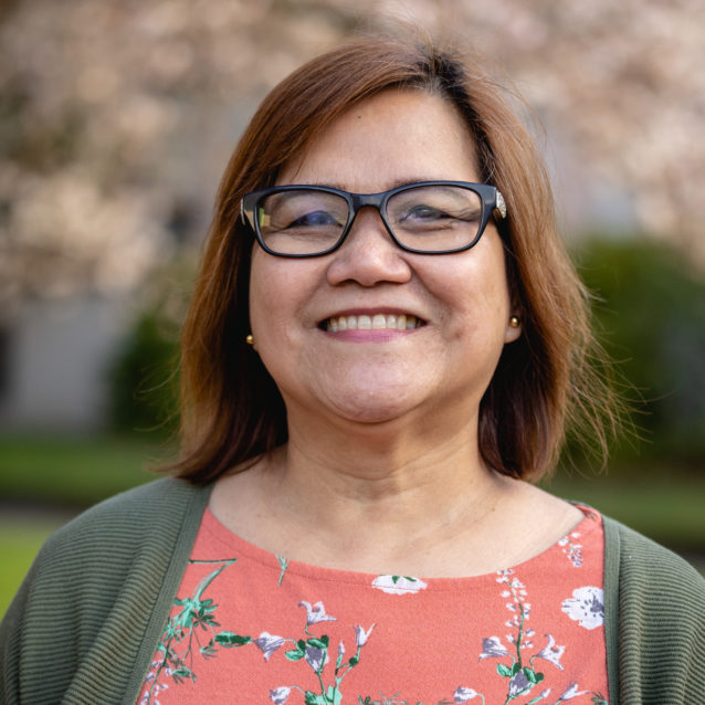 A smiling photo of Lilian Acosta Ma , Health Health. Lilian has short brown hair and is wearing an orange top with a green jacket and glasses.