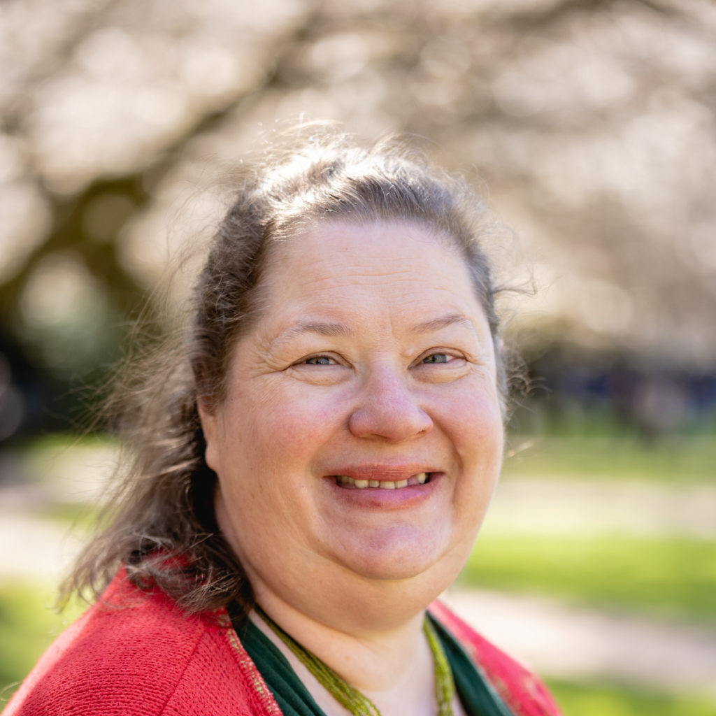 A smiling photo of Ruth Silue Rn , Health Health. Ruth has tied brown hair and is wearing a red top.
