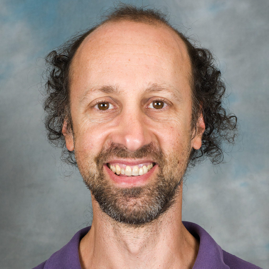 A smiling photo of Howard Levine. Howard has curly black hair and is wearing a purple shirt.