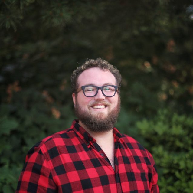 A photo of Jamie Walsh. Jamie wears a red and black checked flannel shirt, has a beard and glasses, and is smiling at the camera.