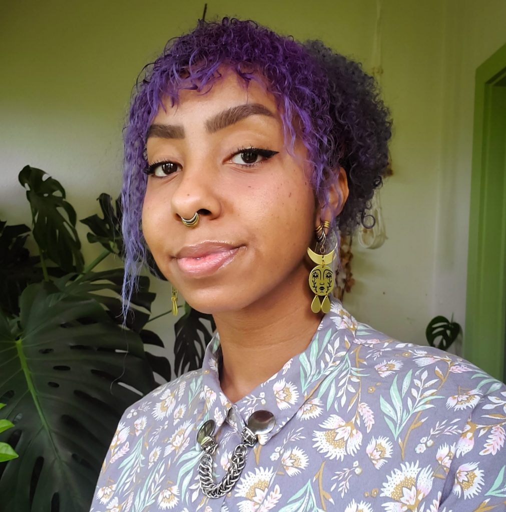 A smiling photo of Davis. Davis has purple curly hair and is wearing a floral purple shirt.