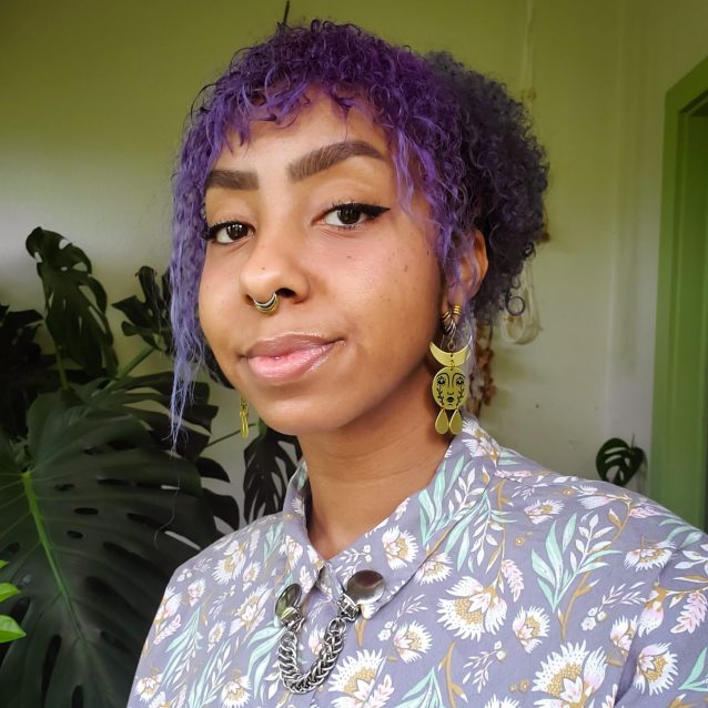 A smiling photo of Davis. Davis has purple curly hair and is wearing a floral purple shirt.