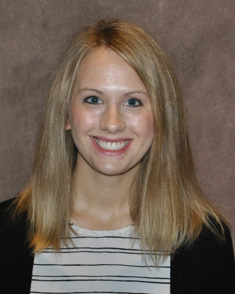 A photo of Kaylin Morrison. Kaylin has medium blonde hair and is wearing a white top with black stripes and a black jacket. She is smiling at the camera in front of a brown background.