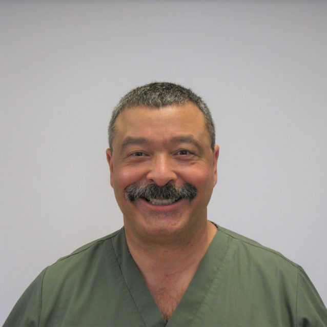 A photo of Delmis Perez. Delmis has short grey/black hair and is wearing a green shirt. He is smiling at the camera in front of a white background.