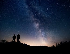 Stars shine in a purple and gold backlit sky as we view two onlookers from the back.