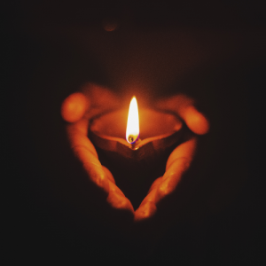 A pair of hands holding a tea light, which shines brightly in a black background.