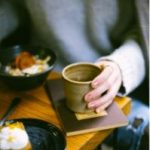 A hand holds a mug sitting on books which sit on a table.