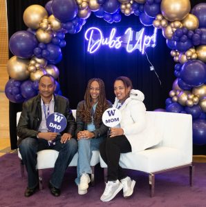A photo of three people of color sitting on a white couch in front of gold and purple balloons and a sign that reads: Dubs Up! A man holds a sign that reads: W Dad and a woman holds a sign that reads: W Mom. A young woman sits between them. They all smile.