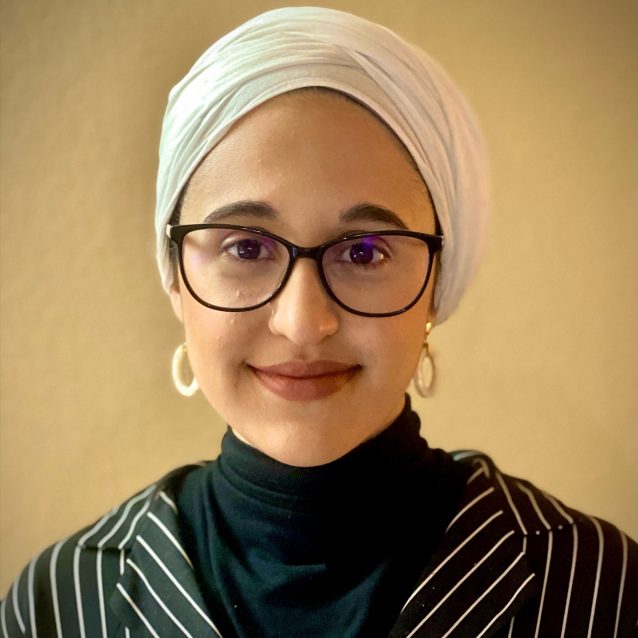 A photo of Faraha Hasan. Faraha is wearing a black turtleneck sweater and a striped blazer. She has on a white wrap-style headscarf, white hoop earrings, and a pair of black-framed glasses. She is smiling into the camera in front of a golden background.