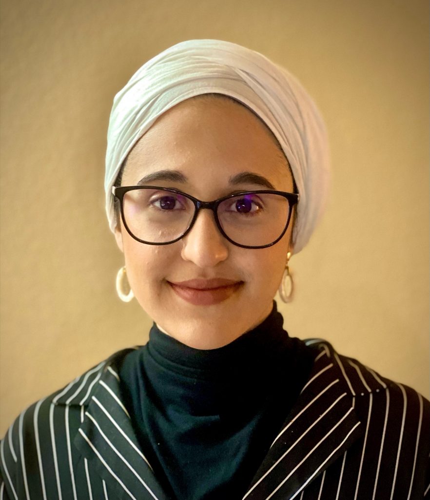 A photo of Faraha Hasan. Faraha is wearing a black turtleneck sweater and a striped blazer. She has on a white wrap-style headscarf, white hoop earrings, and a pair of black-framed glasses. She is smiling into the camera in front of a golden background.