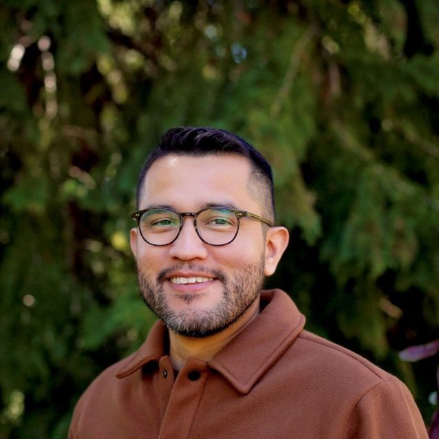 An image of Armando Sanchez in a cinnamon brown shirt wearing glasses with a green nature background.